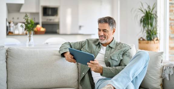 Mann sitzt auf der Couch und ließt lächelnd am Tablet